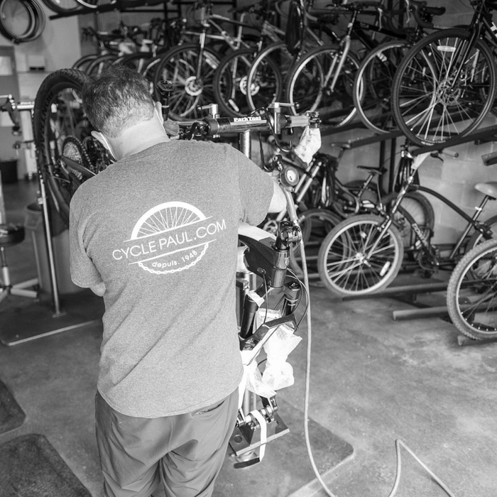 Man repairing a bike, wearing Cycle Paul shirt / Homme réparant un vélo, portant un chandail Cycle Paul