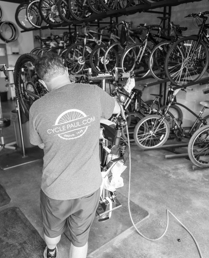 Man repairing a bike, wearing Cycle Paul shirt / Homme réparant un vélo, portant un chandail Cycle Paul
