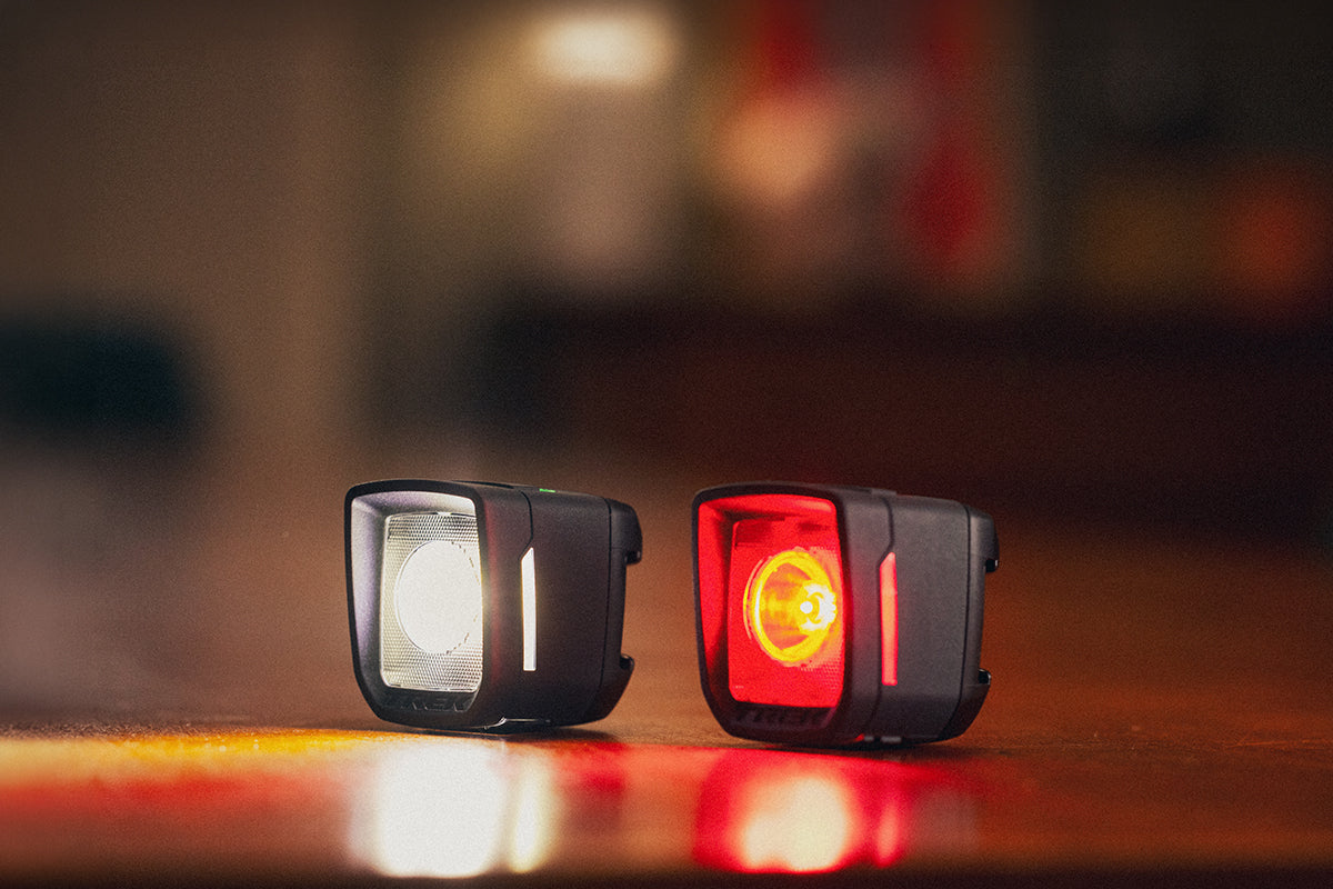 Trek Ion 100 R and Flare R City bicycle lights glowing on a wooden table, featuring a bright white front beam and red rear safety light with a blurred background.