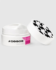 White jar of 'Assos Chamois Crème' with black and white checkered lid on a light gray background