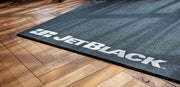 Lifestyle image on hardwood flooring showing the corner of the JetBlack indoor cycling Trainer Mat with white JetBlack logo visible.