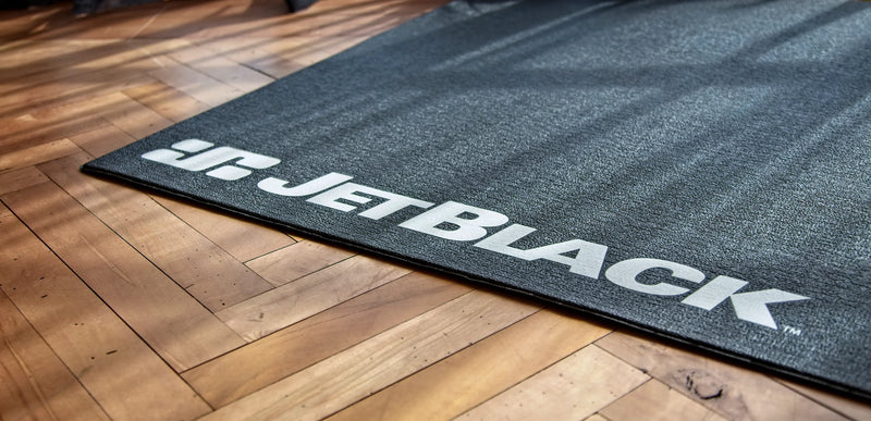 Lifestyle image on hardwood flooring showing the corner of the JetBlack indoor cycling Trainer Mat with white JetBlack logo visible.
