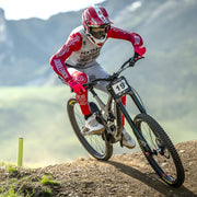 Biker in red and white racing gear riding a mountain bike on a trail with mountains in the background.