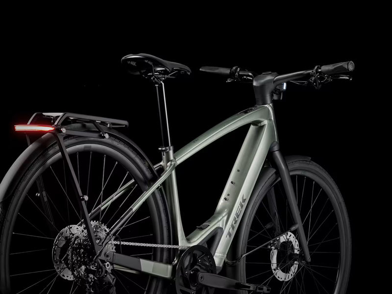 A Trek electric bicycle in a matte sage green frame with black components, shown from a side angle against a solid black background. The brand name 'TREK' is visible on the down tube.