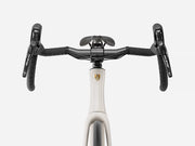 Close-up of a bicycle drop handlebar setup with black grips, black handlebar ends, a black saddle, and a white frame section featuring a gold shield logo, shown against a light gray background.