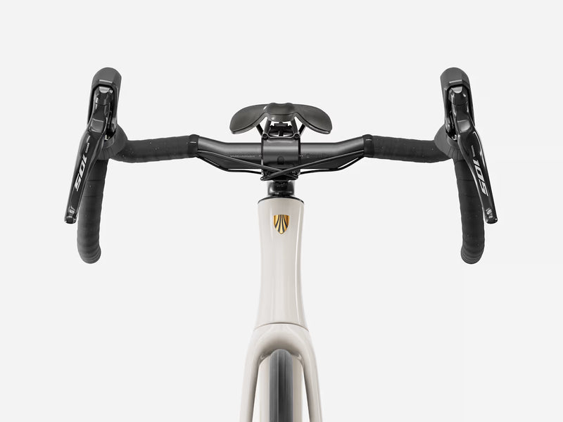 Close-up of a bicycle drop handlebar setup with black grips, black handlebar ends, a black saddle, and a white frame section featuring a gold shield logo, shown against a light gray background.