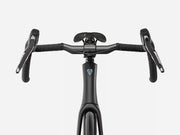 Close-up front view of ablack road endurance bicycle handlebar setup with drop handlebars, black saddle, and a small blue-and-white shield logo on the frame, shown on a light gray background.