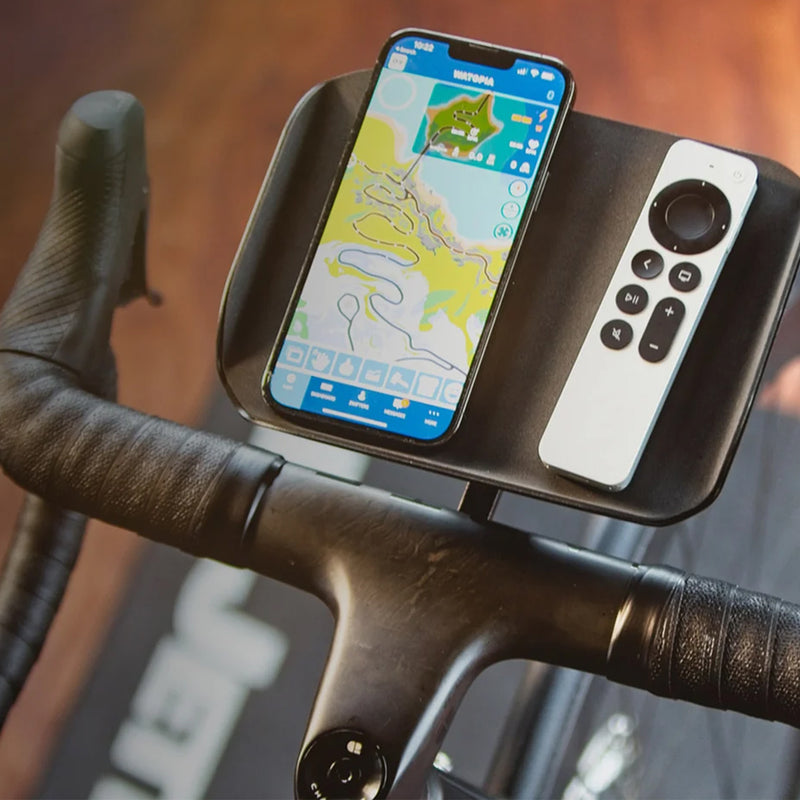 Mounted JetBlack Trainer Tray on a road bike handlebar holding a phone and remote control in a blurred hardwood‑floor living room.