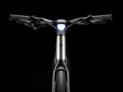 Front view of a modern electric hybrid bicycle with a sleek matte green frame, black handlebars, and a bright illuminated white LED headlight, set against a solid black background.