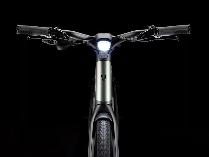 Front view of a modern electric hybrid bicycle with a sleek matte green frame, black handlebars, and a bright illuminated white LED headlight, set against a solid black background.