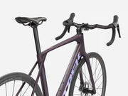 A close-up studio shot of a Trek road bicycle frame and handlebar area, showing a matte dark purple frame with white Trek branding, black saddle, black handlebars, and black wheels against a light gray background.