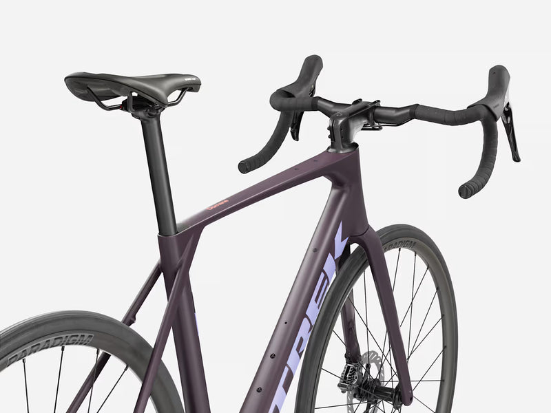 A close-up studio shot of a Trek road bicycle frame and handlebar area, showing a matte dark purple frame with white Trek branding, black saddle, black handlebars, and black wheels against a light gray background.