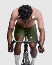 Person wearing green cycling shorts with a logo, riding a stationary bike against a plain background