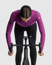 Female cyclist wearing the Assos UMA GT Spring Fall LS Jersey C2 in Purple Alchemy and matching cycling tights, front view on a road bike showing the integrated cable loop and ASSOS logo