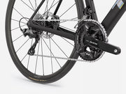 Close-up of a black road bicycle rear wheel and drivetrain, showing the chainring, chain, cassette, rear derailleur, and disc brake on a light gray studio background.