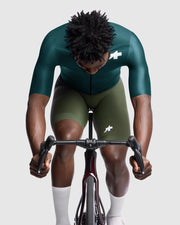 Person wearing a green and olive cycling outfit with a white logo, sitting on a bicycle.
