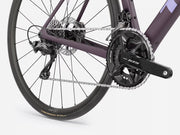 Close-up rear section of a road bicycle showing the dark purple frame, black drivetrain components, chainring, cassette, rear wheel, and disc brake on a light gray studio background.