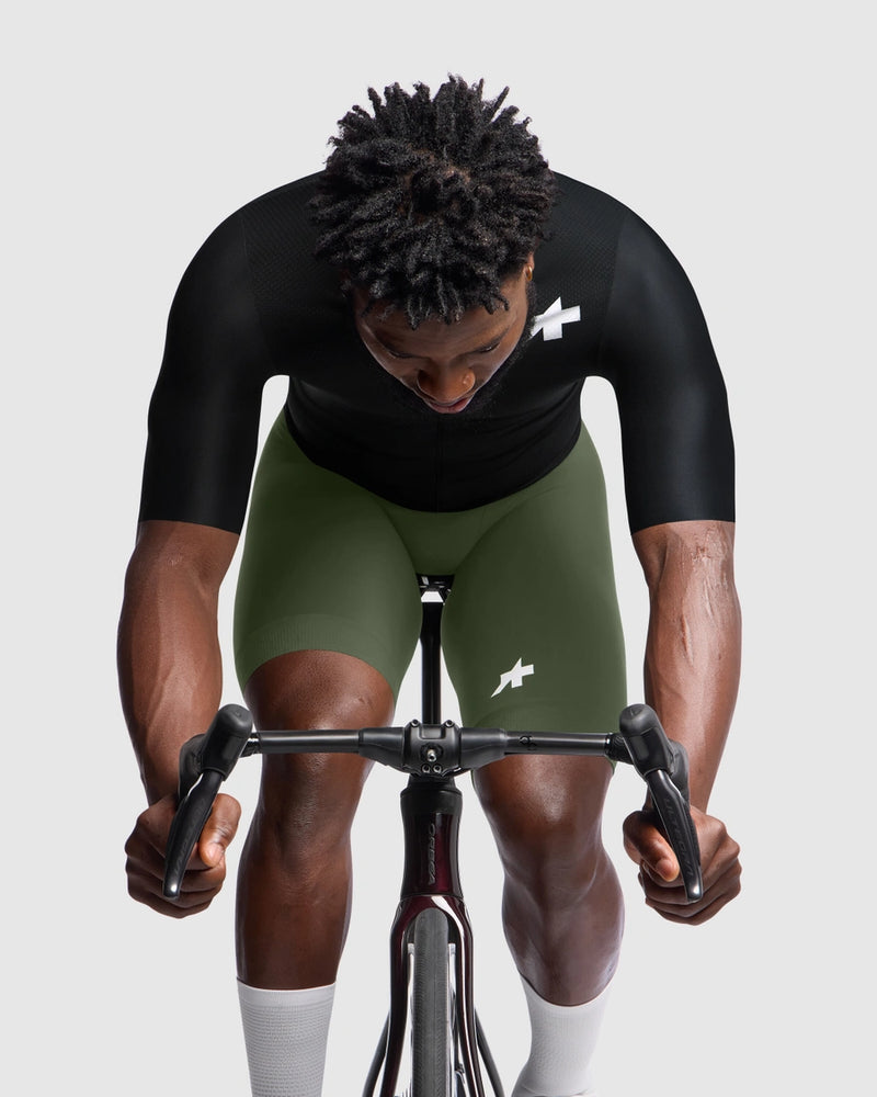 Person wearing a black cycling jersey and green shorts on a stationary bike against a plain background