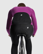Female cyclist wearing the Assos UMA GT Spring Fall LS Jersey C2 in Purple Alchemy, rear view on a road bike showing the black rear pockets and central ASSOS logo
