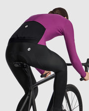 Female cyclist wearing the Assos UMA GT Spring Fall LS Jersey C2 in Purple Alchemy, side view on a road bike showing the jersey fit and lower hem over the cycling tights