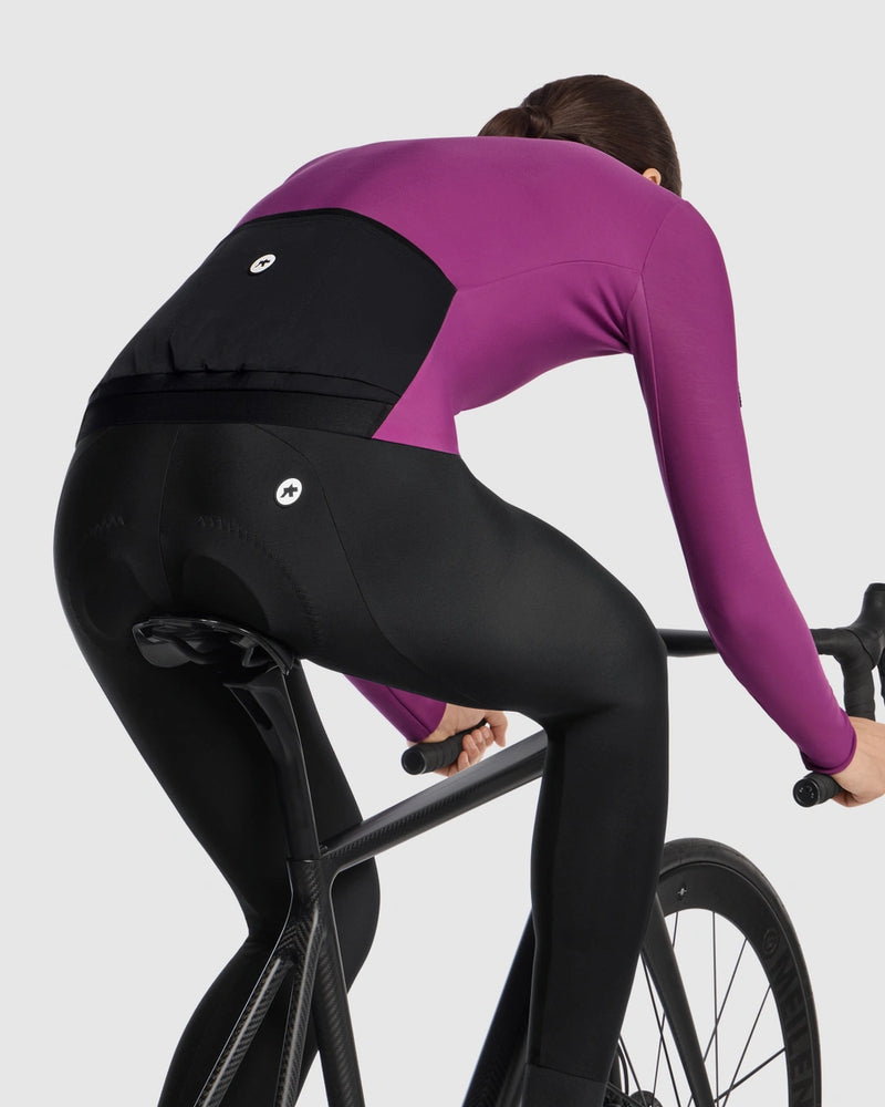 Female cyclist wearing the Assos UMA GT Spring Fall LS Jersey C2 in Purple Alchemy, side view on a road bike showing the jersey fit and lower hem over the cycling tights