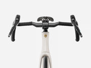 Close-up of an endurance road bicycle drop handlebar with black handlebars and levers attached to a white frame with a gold shield logo, on a light gray background.