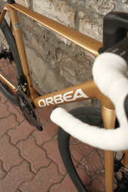 Orca M30ILTD PWR in Metallic Gold finish against a brick wall, closeup of Orbea logo