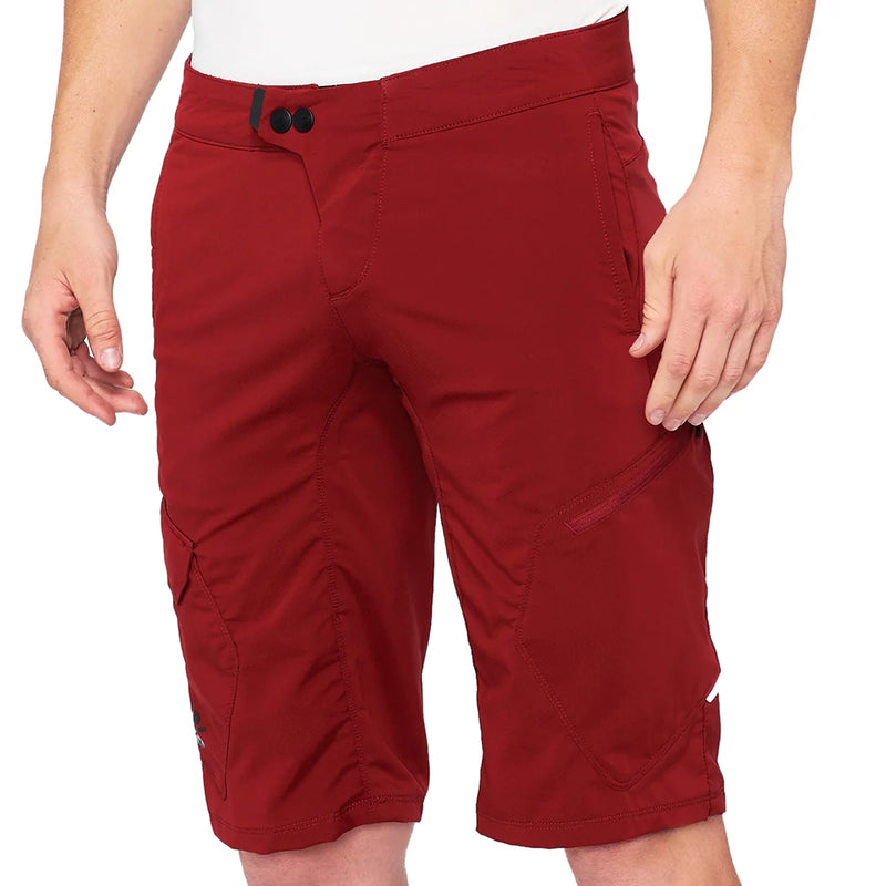 Front product view of brick-red 100% Ridecamp mountain bike shorts featuring snap waist closure, fly detail, precision stitching, and subtle brand logo accents