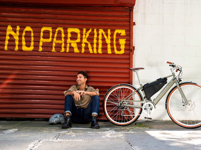 lifestyle image of man, sitting on ground, smiling in front of a garage door that says 'no parking', his helmet on one side and the Electra Loft Go! 7D EQ Step-Over with kickstand on the other.