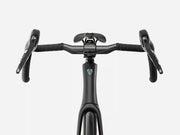Close-up of a black drop handlebar endurance road bicycle setup with a black saddle and black handlebar tape, shown against a light gray studio background. A small blue and white shield logo is visible on the handlebar stem.