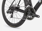 Close-up of a black road bicycle rear wheel and drivetrain, showing the chainring, chain, rear cassette, derailleur, and disc brake on a light gray studio background.