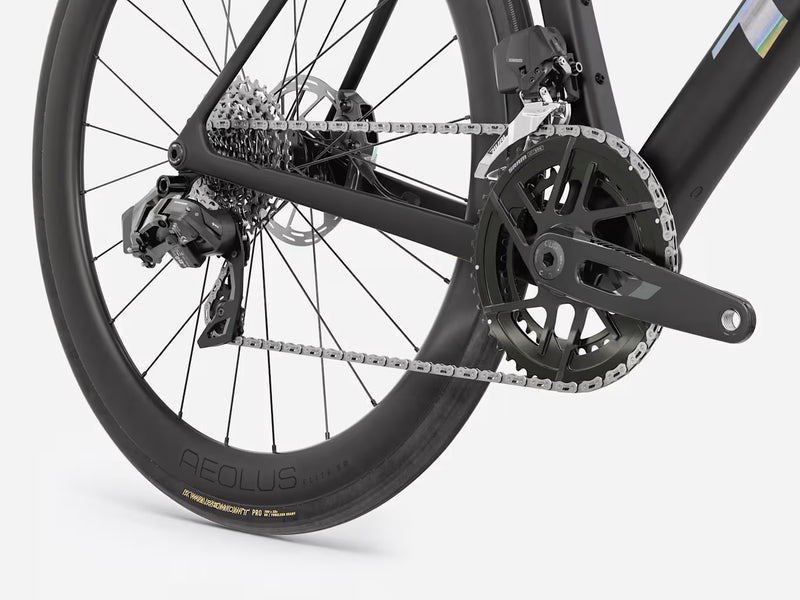 Close-up of a black road bicycle rear wheel and drivetrain, showing the chainring, chain, rear cassette, derailleur, and disc brake on a light gray studio background.
