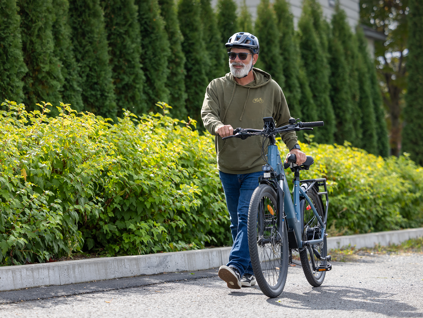 Man with a helmet and sunglasses pushing a bicycle on a path with greenery.
