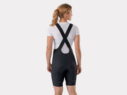 Rear studio view of female model wearing a white undershirt and black Trek Solstice women's bib shorts, displaying the cross-strap configuration and back panel structure