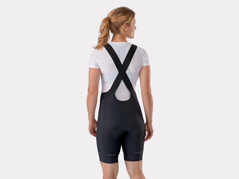 Rear studio view of female model wearing a white undershirt and black Trek Solstice women's bib shorts, displaying the cross-strap configuration and back panel structure
