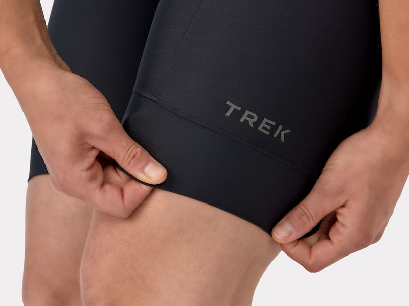 Close-up view of the lower left leg hem on the black Trek Solstice women's bib shorts, showcasing the compressive leg band stitching and prominent Trek logo