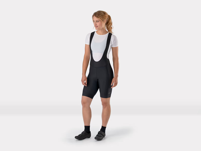 Full-length frontal studio view of female model standing in black Trek Solstice women's cycling bib shorts, showing the complete silhouette from head to toe