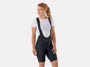 Front studio view of female model wearing a white undershirt and black Trek Solstice women's cycling bib shorts, showing the torso fit, waistband height, and subtle Trek leg branding