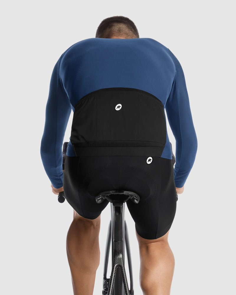 Rear view of a cyclist wearing a blue and black long-sleeve cycling jersey and black cycling shorts with small white circular logos, riding on a light gray background.