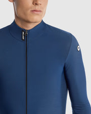 Close-up of a man wearing a blue long-sleeve cycling jersey with a full front zipper and a small circular logo on the left shoulder.
