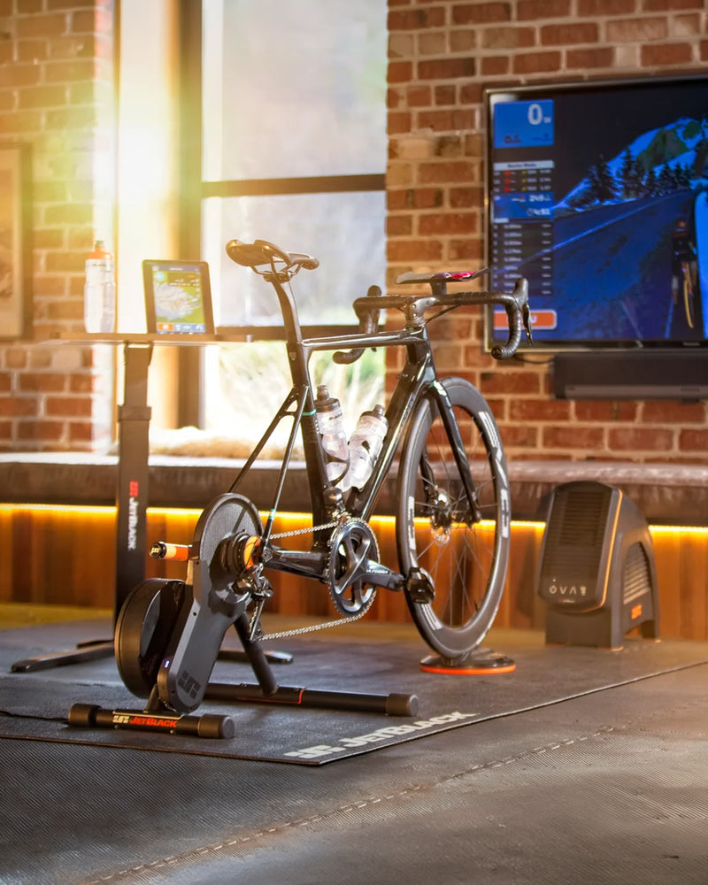 Cyclist training indoors on a JetBlack Victory Smart Trainer with Zwift Cog & Click V2, featuring a road bike, Zwift virtual riding display, JetBlack fan and home workout setup.