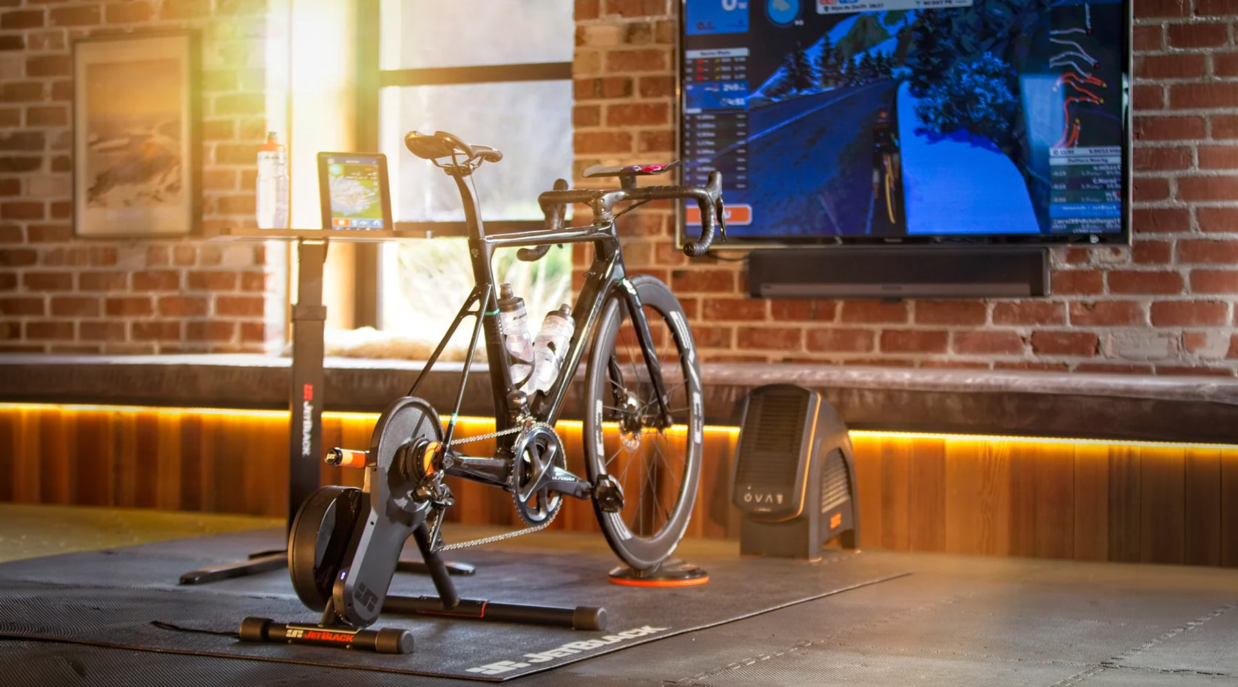 JetBlack Victory Smart Trainer with Zwift Cog & Click V2 set up in a bright home training space with a road bike, trainer mat, JetBlack fan, Zwift display on a television screen, tablet and water bottle beside a sunlit window.