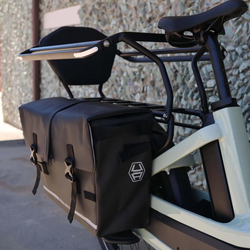 Urban Arrow Breeze Cargo Pannier mounted on the rear of an Urban Arrow Breeze, lifestyle view beside a building showing the front and side of the black pannier with two storage pockets