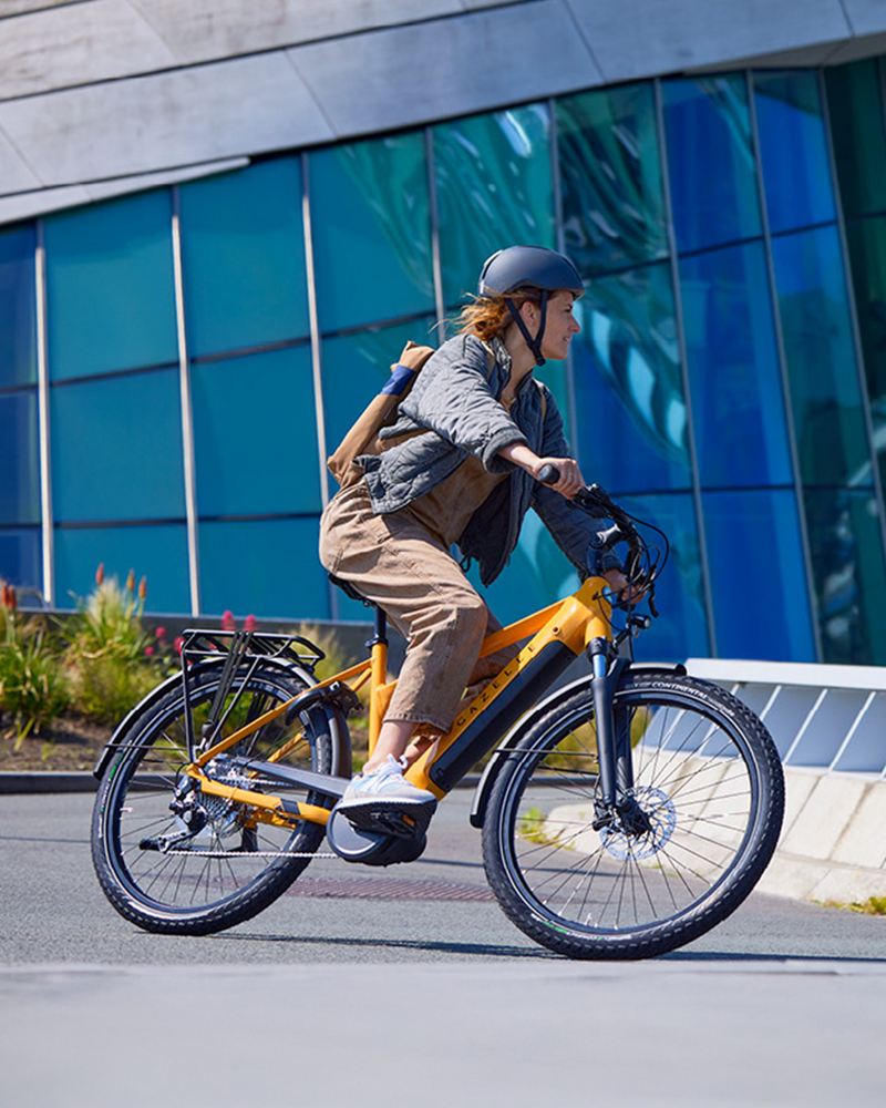 Person riding an electric bike on a city street with modern buildings in the background. / Personne roulant un vélo électrique dans une rue urbaine avec des bâtiments modernes en arrière‑plan.