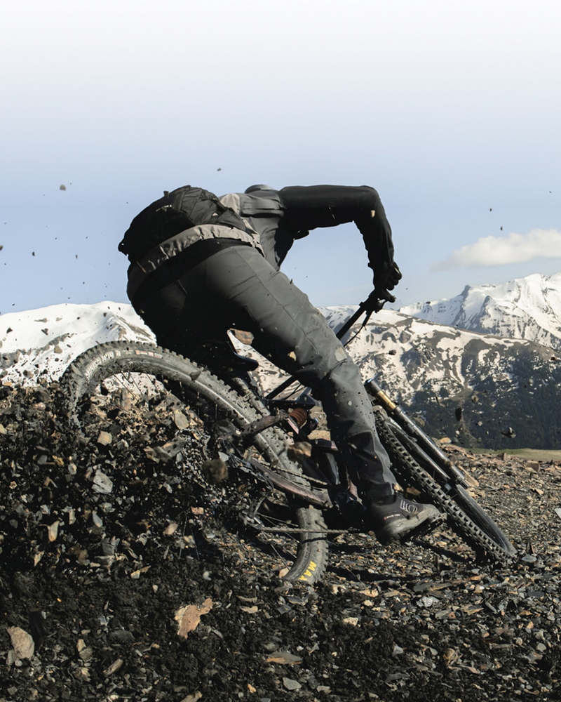 Person riding a mountain bike on a rocky trail with snow-capped mountains in the background. / Personne roulant un vélo de montagne sur un sentier rocheux avec des montagnes enneigées en arrière‑plan.