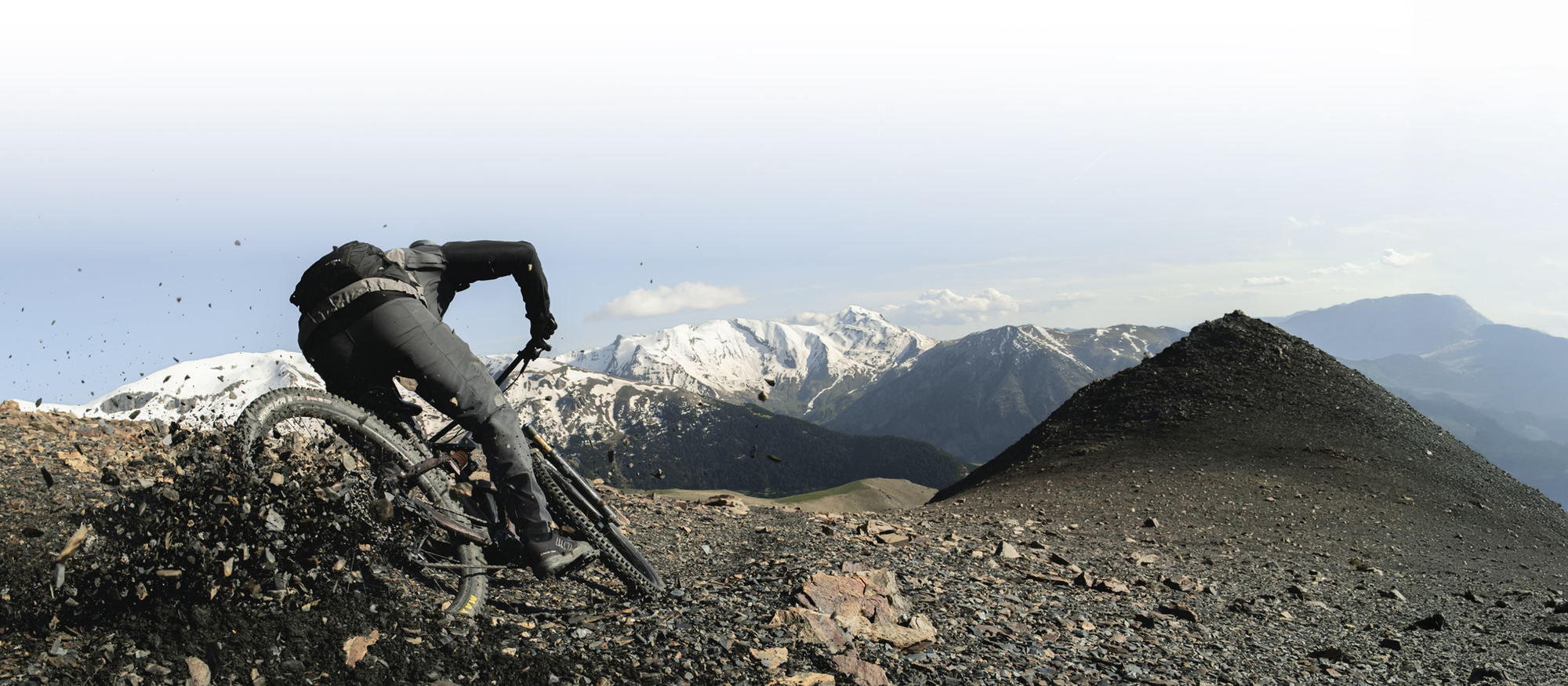 Person riding a mountain bike on a rocky trail with snow-capped mountains in the background. / Personne roulant un vélo de montagne sur un sentier rocheux avec des montagnes enneigées en arrière‑plan.