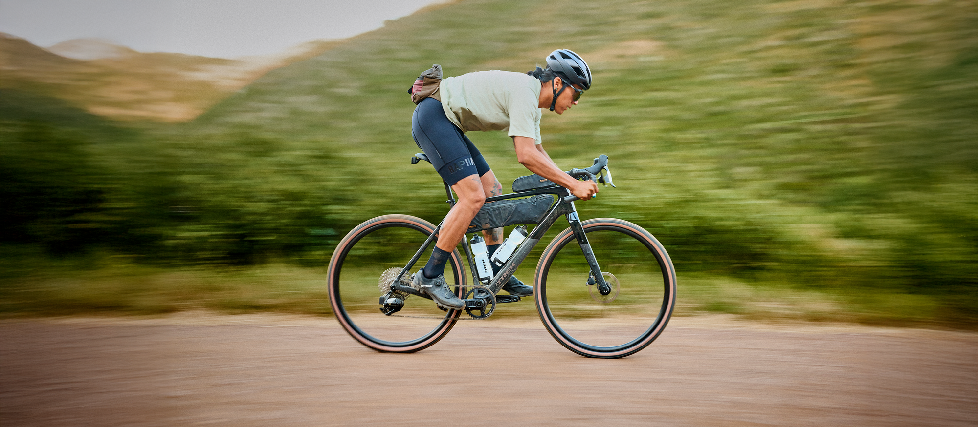 Cyclist riding a road bike at high speed with mountains in the background, motion blur emphasizing speed. / Cycliste roulant à grande vitesse sur un vélo de route avec des montagnes en arrière‑plan, flou de mouvement accentuant la vitesse.